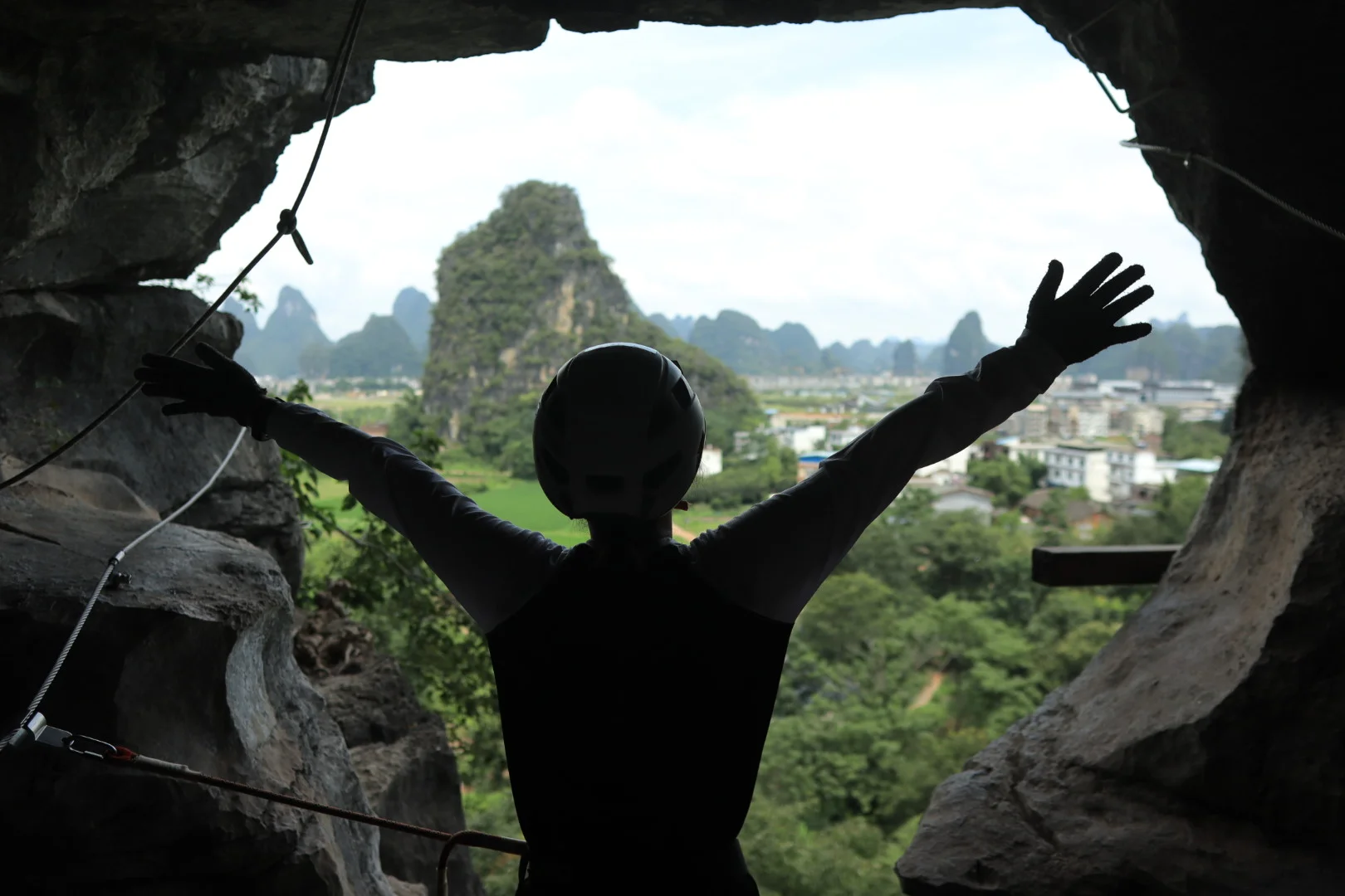 Woman with arms spread wide on a via ferrata cliff edge, Yangshuo karst mountains and valley behind her