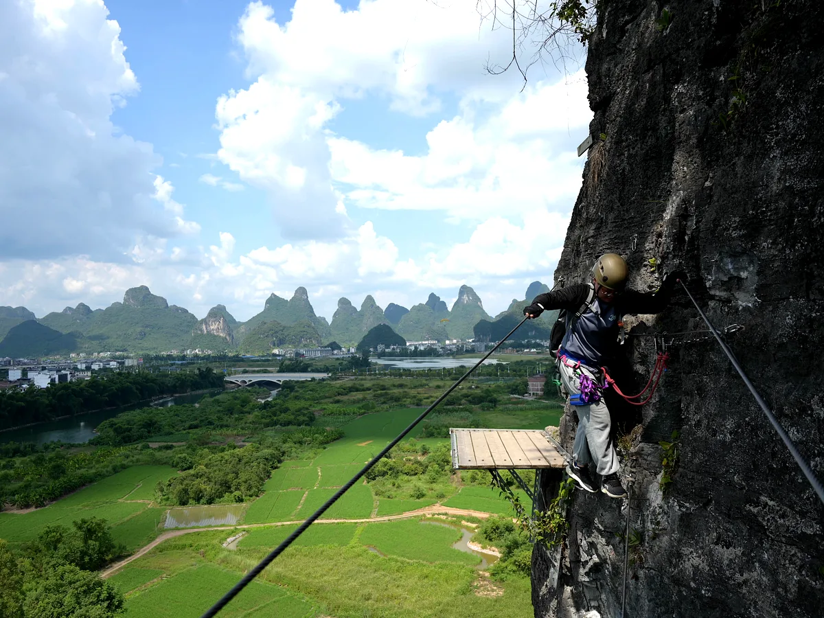 Via ferrata climber in helmet and harness on iron rungs with karst valley, river and village behind, Yangshuo China